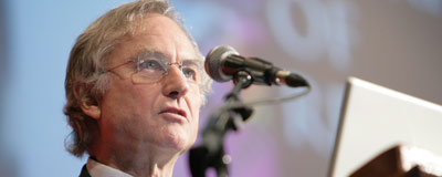 Dawkins mid-lecture. Blurred and in the background is a slide from his lecture and a microphone stand is in the near foreground.