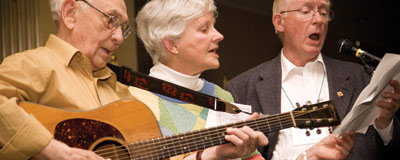 Cropped image of the old-timers going for it. Bleckhorn is playing the guitar in the left foreground. His two friends, slightly behind him, sing passionately into a mic.
