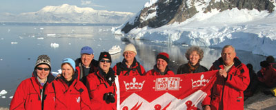 The group stands in matching red outdoor jackets. The four to the right are holding a McGill flag. The eye is drawn to the beautiful sunny vista of ice, open water and craggy mountains behind them. If you look closely at the sea what looks like a huge chunk of ice is actually a cruise ship. Weather looks mild.